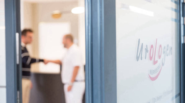 Blick durch die Tür, unscharf dahinter gibt Arzt einem Patienten die Hand.